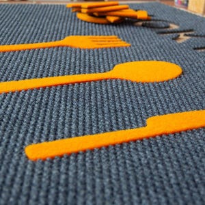 Puede incluir: Primer plano de una superficie rectangular gris texturizada con recortes de fieltro naranja de un tenedor, una cuchara y un cuchillo.