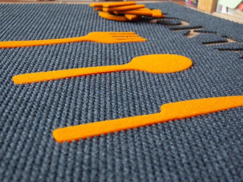 Puede incluir: Primer plano de una superficie rectangular gris texturizada con recortes de fieltro naranja de un tenedor, una cuchara y un cuchillo.