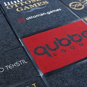 Puede incluir: Primer plano de una variedad de felpudos con diferentes logotipos y dise&ntilde;os. Los felpudos est&aacute;n hechos de tela gris oscuro con bordes negros. Los logotipos incluyen "Ottoman Games", "qubbe LOUNGE" y "BERO TEKSTIL".