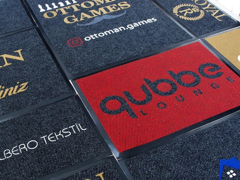 Puede incluir: Primer plano de una variedad de felpudos con diferentes logotipos y dise&ntilde;os. Los felpudos est&aacute;n hechos de tela gris oscuro con bordes negros. Los logotipos incluyen "Ottoman Games", "qubbe LOUNGE" y "BERO TEKSTIL".