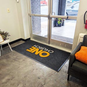 Peut inclure: Un tapis d'entrée rectangulaire noir avec les mots "ONE CHURCH" en blanc et orange. Le tapis est placé devant une porte vitrée, avec un sol en béton gris.