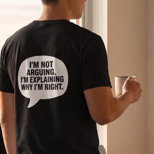 May include: A black short-sleeved t-shirt featuring a white speech bubble graphic on the back with the text: I'M NOT ARGUING. I'M EXPLAINING WHY I'M RIGHT. The shirt is paired with dark trousers, creating a simple and casual outfit for daily wear.