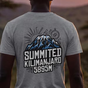 May include: A light grey t-shirt featuring a graphic on the back with a stylised mountain peak, a sun, and a compass. The text reads "SUMMITED KILIMANJARO 5895M" in bold white letters. The design uses blue, white, and black ink on the fabric.