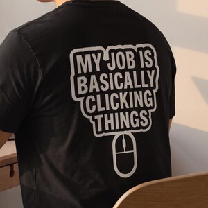 Può includere: Una t-shirt nera a maniche corte con testo bianco sul retro che recita MY JOB IS BASICALLY CLICKING THINGS sopra la grafica di un mouse per computer. La maglietta è indossata da una persona seduta a una scrivania con un portatile.