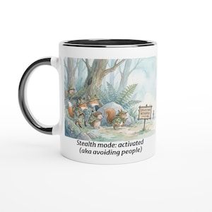 May include: White ceramic mug with a black rim and handle. The mug features a whimsical illustration of woodland creatures in camouflage, with the text "Stealth mode: activated (aka avoiding people)".