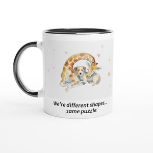 Pode incluir: Caneca de cerâmica branca com alça e borda pretas. A caneca apresenta uma ilustração em aquarela de uma girafa e um cachorro se abraçando. O texto na caneca diz: "We're different shapes... same puzzle."