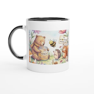 Puede incluir: Taza de cerámica blanca con asa y borde negros. La taza presenta una ilustración caprichosa de un oso con un tarro de miel, una abeja, un erizo, una ardilla y el texto "I'm un-bee-lievably missing you."