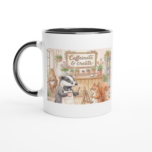 Op de afbeelding: Witte keramische mok met een zwarte rand en oor. De mok heeft een aquarel illustratie van een das en een eekhoorn in een kunststudio met de tekst "Caffeinate & create". De achtergrond bevat planten en een houten toonbank.