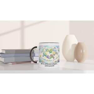 Pode incluir: Caneca de cerâmica branca com alça e borda pretas. A caneca apresenta uma ilustração colorida de várias formas e criaturas formando um coração, com o texto "We're different shapes... same puzzle".