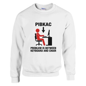 Może przedstawiać: Biała bluza z napisem "PIBKAC" nad grafiką przedstawiającą osobę siedzącą przy komputerze, ze strzałką w dół. Poniżej grafiki znajduje się napis "PROBLEM IS BETWEEN KEYBOARD AND CHAIR."