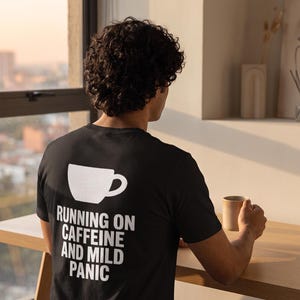 Könnte beinhalten: Ein schwarzes Kurzarm-T-Shirt mit einer weißen Grafik einer Kaffeetasse auf der Rückseite. Unter der Tasse steht der Text "RUNNING ON CAFFEINE AND MILD PANIC" in fetten, weißen Großbuchstaben. Das T-Shirt besteht aus einfarbigem schwarzem Stoff.