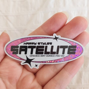 Op de afbeelding: Een ovale sticker in roze en wit met glitter. De sticker heeft de tekst "HARRY STYLES SATELLITE SPINNING OUT WAITING FOR YA" in zwart.