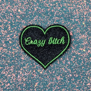 May include: A black heart-shaped patch with green stitching and the text "Crazy Bitch" on a glittery blue background.
