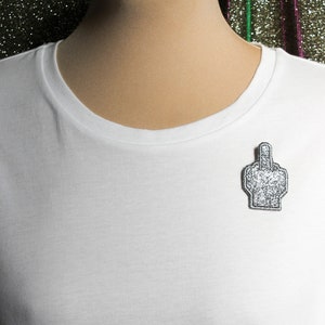 May include: White t-shirt with a silver glitter middle finger pin.
