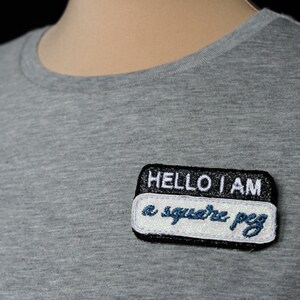 Peut inclure: Un t-shirt gris à col rond avec un écusson noir et blanc. L'écusson indique "HELLO I AM a square peg" en lettres blanches et bleues. L'écusson est rectangulaire avec des coins arrondis.