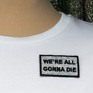 Può includere: Maglietta bianca con una toppa di glitter argentata che dice "WE'RE ALL GONNA DIE" in testo nero.