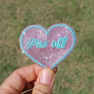 May include: Pink glitter heart-shaped patch with a turquoise blue outline and the text "Piss off" embroidered in turquoise blue.