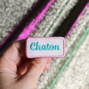 May include: A pink and white iron-on patch with the word "Chaton" embroidered in turquoise blue.