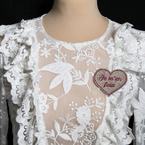 Può includere: Top in pizzo bianco con maniche lunghe e scollo a volant. Una toppa a forma di cuore con il testo "Je m'en fous" in lettere rosa è attaccata alla parte anteriore del top.