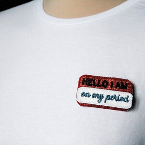 以下が含まれることがあります： 「HELLO I AM on my period」と書かれた赤と白のネームタグが付いた白いTシャツです。