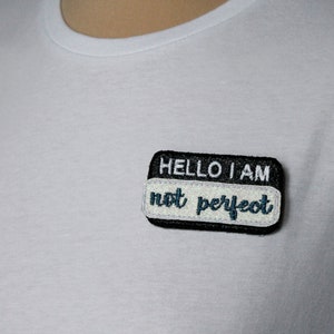 May include: White t-shirt with a black and white patch that reads "HELLO I AM not perfect" in a teal font. The patch is rectangular with rounded corners and is sewn onto the shirt.