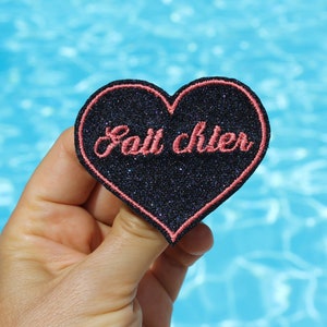 Può includere: Una toppa a forma di cuore con sfondo glitterato nero e bordo rosa. La toppa presenta la frase ricamata "Fait chier" in corsivo rosa. Lo sfondo è una superficie d'acqua azzurra.