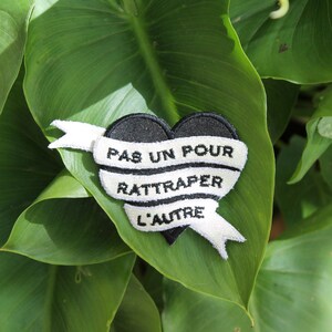 May include: Black heart-shaped patch with white ribbon and the text "PAS UN POUR RATTRAPER L'AUTRE" on a green leafy background.