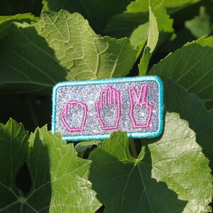 May include: A rectangular patch with a silver glitter background and a teal border. The patch features pink outlines of the hand gestures for rock, paper, and scissors. The patch is set against a backdrop of green leaves.