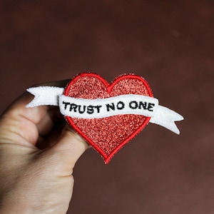Könnte beinhalten: Rotes Glitzerherz mit einem weißen Banner, auf dem in schwarzer Stickerei "TRUST NO ONE" steht.