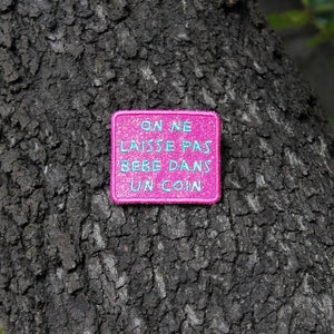 Könnte beinhalten: Rosa Stoff-Patch mit dem Text "On ne laisse pas bebe dans un coin" in blauer Schrift. Der Patch ist an einem Baumstamm befestigt.