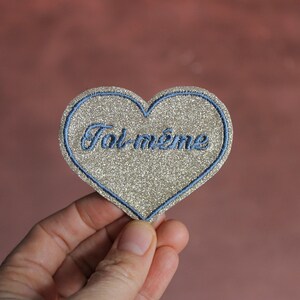 May include: A heart-shaped patch with a silver glitter design and a blue border. The French text "Toi même" is embroidered in blue script across the center. The patch is held against a pink background.