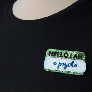 May include: A black t-shirt with a patch that reads "HELLO I AM a psycho." The patch has a green glitter border and a white background with blue text. The shirt is plain and dark.
