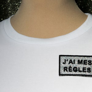 Brosch "JAG FÅR MIN MENS", silvertyg med glittrande regnbågspaljetter, broderat meddelande på franska, svart trådkontur, tillverkad i Frankrike