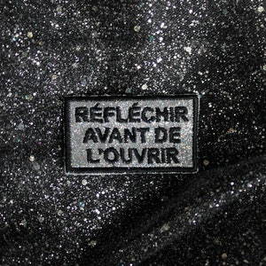 Peut inclure: Un patch rectangulaire noir et argent avec le texte "Réfléchir avant de l'ouvrir" sur un fond noir pailleté.