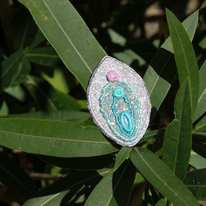 Op de afbeelding: Een zilveren glitterpatch met een geborduurd ontwerp in turquoise en roze. De patch heeft de vorm van een traan en heeft een reliëf, textuur.