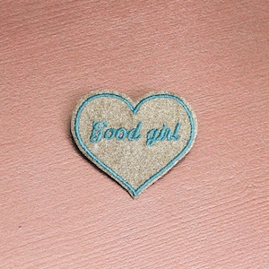 May include: A heart-shaped patch with the words "Good girl" embroidered in teal thread. The patch has a tan background and a teal border. The patch is likely for clothing or accessories.
