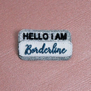May include: A rectangular name tag with the text "HELLO I AM" in black and "Borderline" in blue script. The tag has a silver glitter border and a white background for the name.