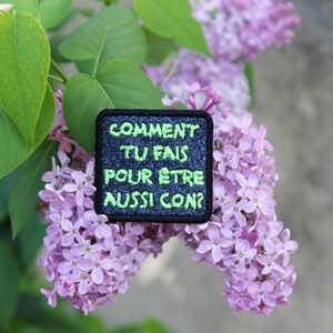May include: A black patch with green glitter text that reads "COMMENT TU FAIS POUR ÊTRE AUSSI CON?"  The patch is on a branch of purple lilac flowers.