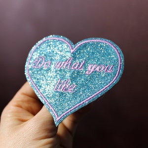 May include: A heart-shaped patch with a glittery blue surface and a pink outline. The patch features the phrase "Do what you like" in pink script. The patch is held in a hand against a dark background.