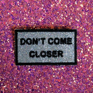 Puede incluir: Un parche rectangular con las palabras "DON'T COME CLOSER" en letras negras sobre un fondo plateado brillante, enmarcado por un borde negro. El fondo es un brillante rosa brillante.