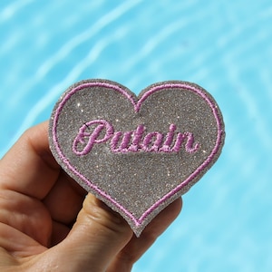 Könnte beinhalten: Ein herzförmiger Aufnäher mit einer glitzernden silbernen Oberfläche und einem rosa Rand. Das Wort "Putain" ist in rosa Schrift über die Mitte gestickt. Der Aufnäher wird vor einem blauen Wasserhintergrund gehalten.
