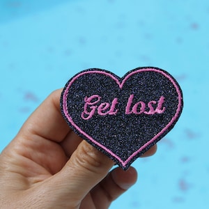 Könnte beinhalten: Ein herzförmiger, gestickter Aufnäher mit dem Schriftzug "Get lost" in rosa. Der Aufnäher hat einen dunkelblauen Glitzerhintergrund und einen rosa Rand. Der Aufnäher wird vor einem hellblauen Hintergrund gehalten.