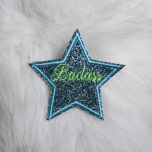 Peut inclure: Écusson en forme d'étoile pailletée bleu sarcelle avec une bordure bleu clair et le mot "Badass" brodé en vert. L'écusson est posé sur un fond blanc et duveteux.