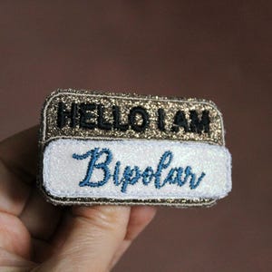 May include: A gold glitter name tag with the text "HELLO I AM" and "Bipolar" in blue cursive lettering.