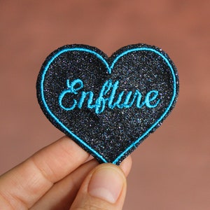 Puede incluir: Parche negro en forma de corazón con purpurina azul y texto bordado en azul que dice "Enflure".