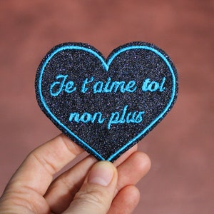 May include: A black heart-shaped patch with blue glitter and blue embroidered text that reads "Je t'aime toi non plus".