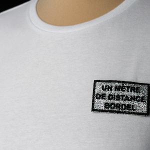 Könnte beinhalten: Weißes T-Shirt mit einem silbernen Glitzer-Patch, auf dem "UN METRE DE DISTANCE BORDEL!" steht.