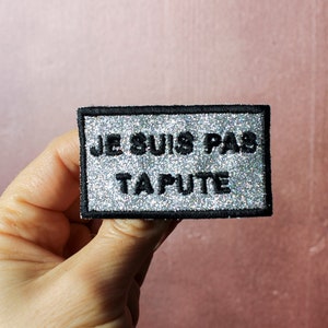 May include: A silver glitter iron-on patch with black text that reads "JE SUIS PAS TA PUTE".