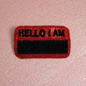 Könnte beinhalten: Ein rechteckiger Namensschild-Aufnäher mit abgerundeten Ecken. Der obere Teil ist rot mit Glitzer und den schwarzen Worten "HELLO I AM". Der untere Teil ist schwarz und leer.
