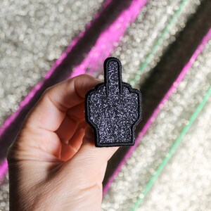 May include: A black glitter patch in the shape of a hand giving the middle finger. The patch has a black border and is held up against a sparkly background.